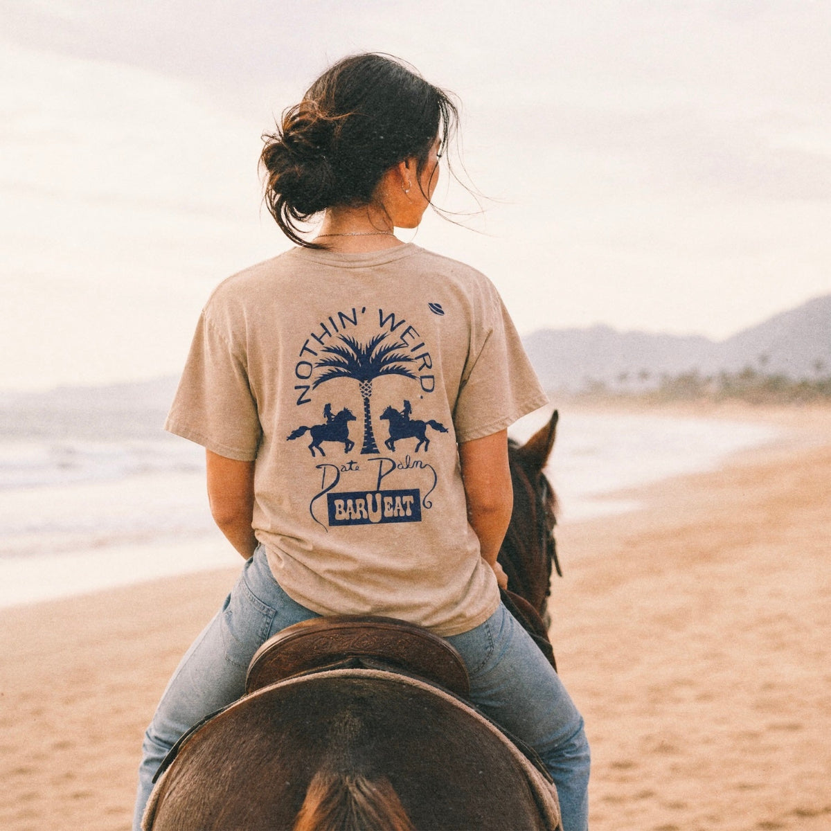 woman on the beach on horseback wearing a graphic tee with a palm tree on it