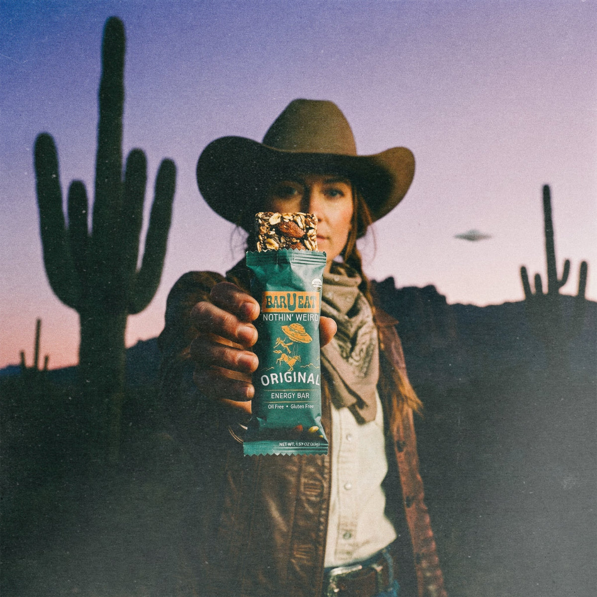 Person in cowboy hat holding a barueat original energy bar with cacti and UFO in the background