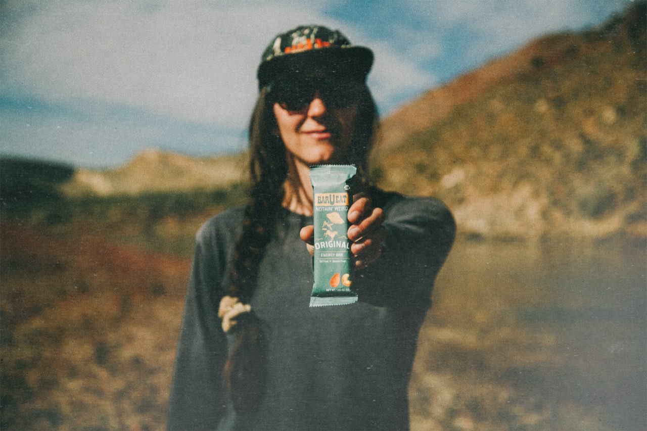 A hiker holding out a Bar U Eat Original energy bar beside a mountain river